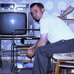 Ein Mann sitzt auf einem Stuhl vor einem Fernseher. Hinter ihm steht eine Frau. Neben ihm steht ein Dinett. 