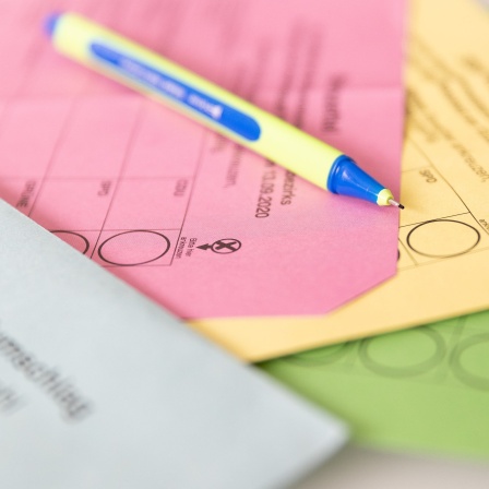 Verschiedene Stimmzettel und ein Stimmzettelumschlag für die Briefwahl liegen auf einem Tisch zusammen mit einem Stift.