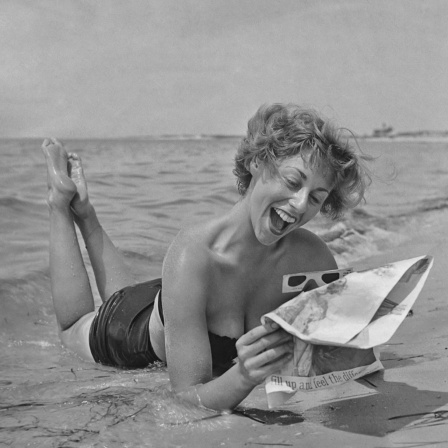 Schwarzweißfoto einer Frau, die im Bikini am Strand liegt und eine Zeitung liest.