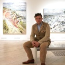 Christopher Lehmpfuhl sitzt auf einer Bank, hinter ihm hängen drei seiner Bilder an der Wand