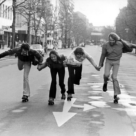 Fünf Menschen halten sich an den Händen und fahren Rollschuh auf einer leeren Straße.