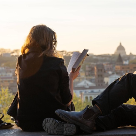 Eine junge Frau und ein junger Mann sitzen Bücher lesend auf einer Mauer im Orangengarten in Rom, mit dem Petersdom und Sonnenuntergang im Hintergrund