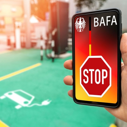 Ein Smartphone zeigt das Wort "BAFA", darunter ein Stopschild. Im Hintergrund ist ein Parkplatz für Elektro-Autos zu sehen, auf dem man das Auto abstellt, um dann die Batterie des Autos laden zu können.