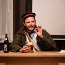 Im Bild aus der Inszenierung "saufen fechten heidelberg" sitzt ein Mann mit Uniformmütze an einem Tisch, hält ein Bierglas in der einen Hand und formt mit der anderen einen Ring aus Daumen und Zeigefinger.