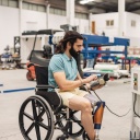 Ein Mann mit Beinprothese sitzt in einem Rollstuhl und bedient eine Maschine in einer Industriehalle.