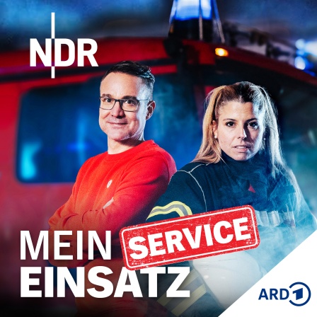 Ein Mann in rotem Pullover und eine Frau in Feuerwehruniform mit Helm stehen vor einem roten Feuerwehrauto mit Blaulicht und Rauch im Hintergrund.