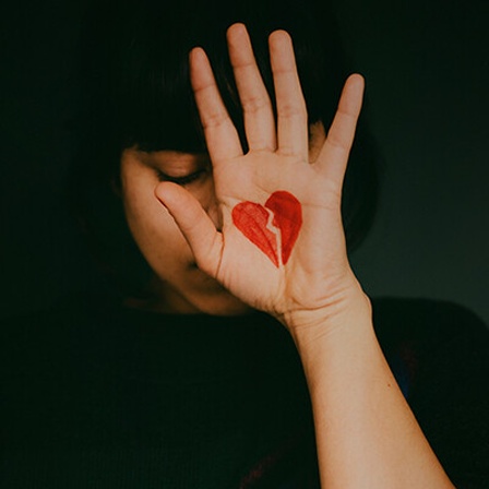 Eine Frau hält ihre Hand hoch vor ihr Gesicht, auf die Handfläche ist ein gebrochenes Herz gemalt.