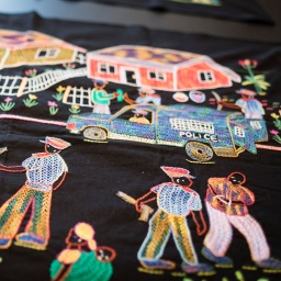 Kunstwerke des Artivism Atrocity Prevention Pavillons des Auschwitz Instituts Peace and Reconciliation: Stickerei zeigt Polizeiautos, die durch eine Nachbarschaft mit spielenden Kindern und sich unterhaltenden Erwachsenen fahren.