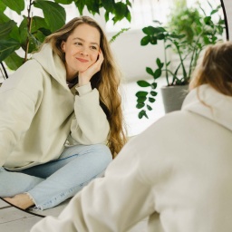 Junge Frau sitzt, umgeben von Pflanzen, vor einem Spiegel und lächelt