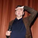 Christian Petzold steht vor einem Vorhang auf einer Bühne und schaut mit einer Handfläche an die Stirn gelegt in die Ferne. 