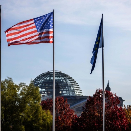 Die Flaggen Deutschlands, der USA und der EU wehen im Wind, im Hintergrund der Reichstag in Berlin. 