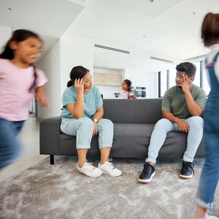 Ein Paar sitzt auf dem Sofa und schaut sich fragend an, während die Töchter im Zimmer herumrennen. 