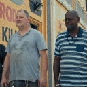 Drei Männer um die 50, ein Schwarzer und zwei Weiße, stehen in T-Shirts mit ernsten Gesichtern vor einer Backsteinfassade.