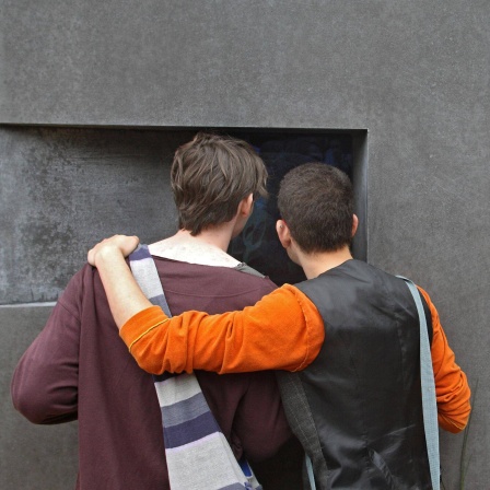 Zwei Männer stehen Arm in Arm vor dem Denkmal für die im Nationalsozialismus verfolgten Homosexuellen in Berlin. 