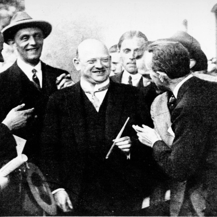 Eine Schwarz-Weiß-Fotografie zeigt eine Reihe lachender Männer auf einen kahlköpfigen Mann mit langer Zigarre zeigend.