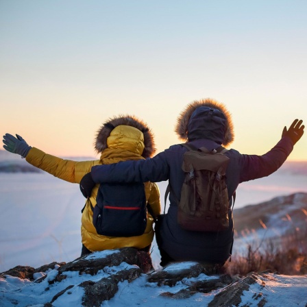 Zwei Wanderer strecken zum Sonnenuntergang in einer verschneiten Landschaft die Arme aus (Bild: picture alliance/Zoonar/Dasha Petrenko)