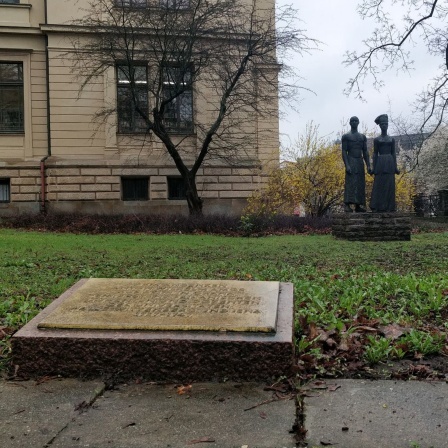 Im Vordergrund eine Gedenktafel auf dem Boden, rundherum eine Wiese, im Hintergrund zwei Statuen und ein Gebäude