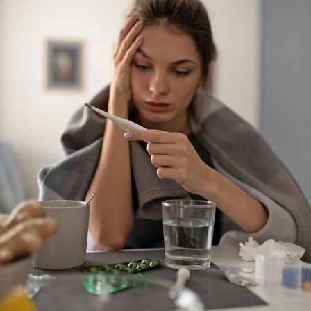 Junge Frau mit einer Erkältung schaut auf ein Fieberthermometer.