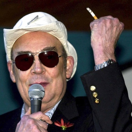 Der Hippie Chronist Hunter S. Thompson trägt Sonnenbrille und Hut, in der rechten Hand ein Mikrofo, in der linken eine Zigarette.