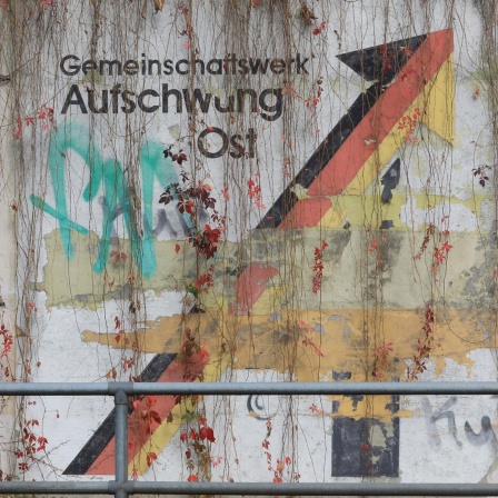 Auf einer abgeblätterten Mauer ist der Aufdruck "Gemeinschaftswerk Aufschwung Ost" mit Graffitis übersprüht.
