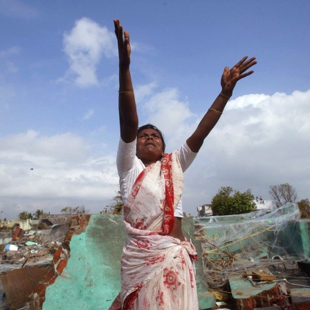 Eine indische Fischerfrau reckt hilfesuchend ihre Arme in die Höhe, um einen Hubschrauber zu erreichen. Hinter ihr die Trümmer nach dem Tsunami 2004