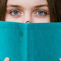Eine Frau mit blauen Augen schaut etwas ängstlich hinter einem Buch mit blauem Umschlag hervor.