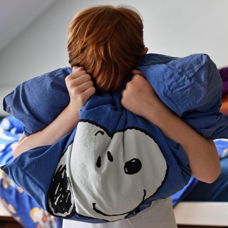 Ein Junge steht mit seinem Kuscheltier und einem Kopfkissen in den Händen vor seinem Bett im Kinderzimmer (gestellte Szene).