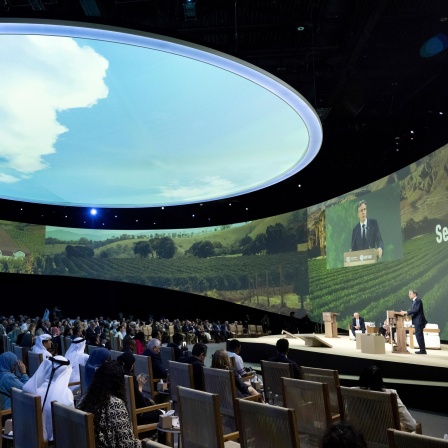 In einer Halle sitzen mehrere Vertreter im Publikum während auf der Bühne gesprochen wird. Die Decke der Halle ist mit einem riesigen Kreis perforiert sodass der Himmel zu sehen ist.