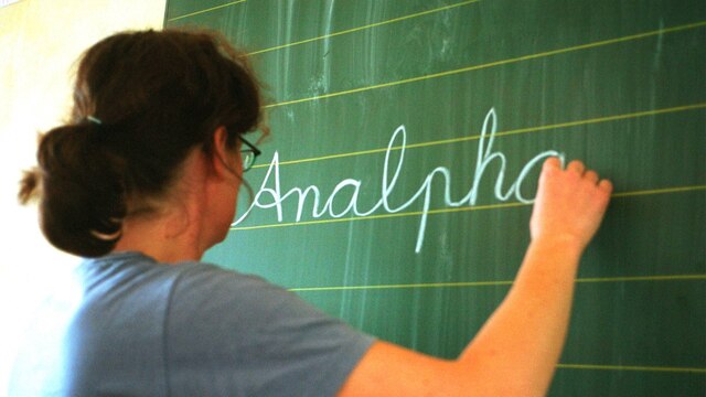 Eine Frau steht an der Tafel und schreibt mit Kreide das Wort "Analphabetismus" an