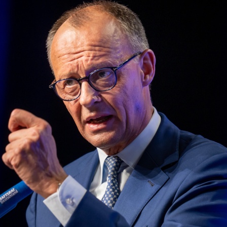 Friedrich Merz  in einem blauen Anzug mit passender Krawatte zu sehen. Er trägt eine runde Brille und hat kurzes, lichtes Haar. Die Person spricht offensichtlich in einem öffentlichen Rahmen, vermutlich auf einer Veranstaltung oder Konferenz.