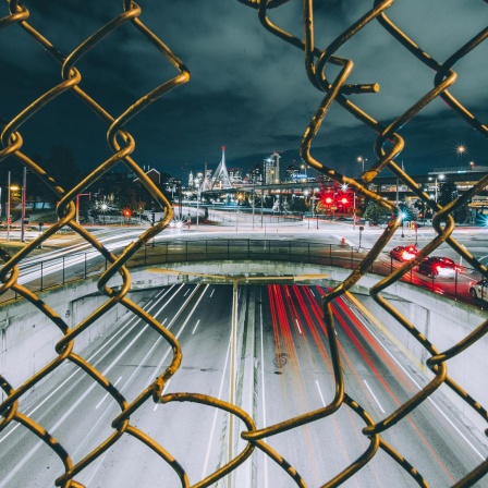Blick durch einen Maschendrahtzaun auf eine dramatisch beleuchtete Stadt bei Nacht