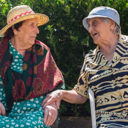 Zwei ältere Frauen mit Sonnehüten sitzen auf Stühlen und unterhalten sich
