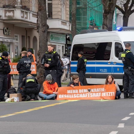Aktivisten der Gruppe "Letzte Generation" haben sich auf der Neuen Kantstraße auf einer Kreuzung festgeklebt. Die Polizei sichert den Bereich.