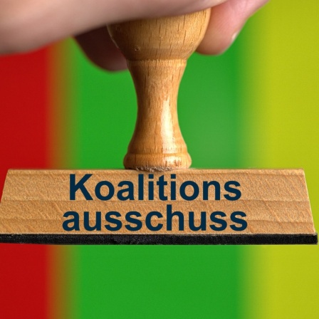 Ein symbolischer Holzstempel mit der Aufschrift "Koalitionsausschuss", gehalten von einer Hand, vor den Farben der Ampel-Koalition. 