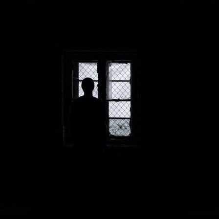 Eine Frau steht als Silhouette im Dunkeln vor vergitterten Fenstern und schaut hinaus