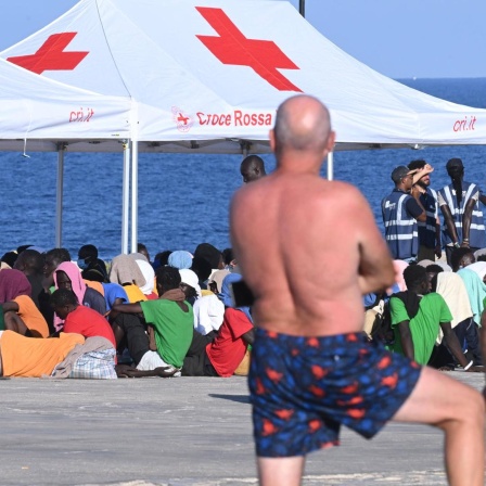 Geflüchtete sitzen unter einem Zelt des Roten Kreuzes im italienischen Lampedusa. Im Vordergrund steht ein Mann in Badehose und beobachtet sie. 