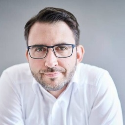 Portrait von Wirtschaftspsychologe Carsten Schermuly: Dunkelhaariger Mann mit Brille und kurzem Bart im weißen Hemd vor grauer Wand