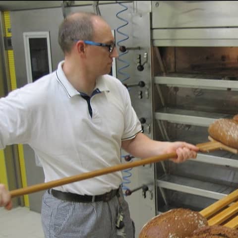 Zwei Bäcker in weißen Oberteilen und schwarz-weiß karierten Hosen stehen vor einem Industriebackofen in der JVA Tonna und heben frisch gebackene Brote heraus