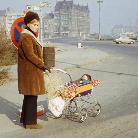 Eine Frau mit Kinderwagen, in dem ein Baby schläft, steht mit nachdenklichem Blick an einer Straßenecke im Berlin der Siebzigerjahre. Die Spuren des Kriegs sind noch im Stadtbild zu erkennen.