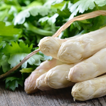 Frischer weißer Spargel und ein Bund Petersilie liegen auf einem Holztisch.