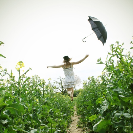 Eine junge Frau in einem Sommerkleid läuft durch ein Feld und wirft einen Regenschirm in die Luft.