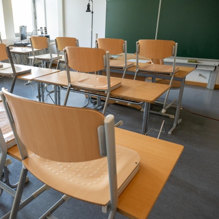 Ein leeres Klassenzimmer mit hochgestellten Stühlen und einer grünen Tafel im Hintergrund.