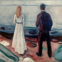 Ein Paar steht am Strand und schaut auf's Meer. Der Mann steht ein wenig hinter der Frau und scheint ebenfalls auf sie zu blicken.