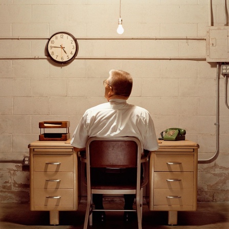 Ein Mann sitzt vor einer eintönigen Wand und schaut auf die Uhr, die schräg links über ihm hängt.