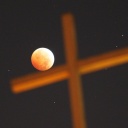 Ein religiöses Kreuz vor dem Nachhimmel, während einer totalen Mondfinsternis. Der rot erleuchtete Mond scheint das Kreuz zu berühren.