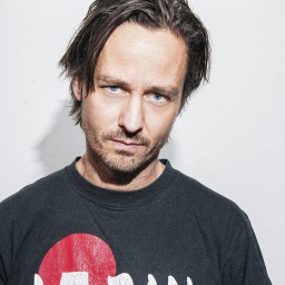 Ein Mann mittleren Alters mit dunklem T-Shirt und rot, weißer Aufschrift schaut mit tiefem Blick in die Kamera.