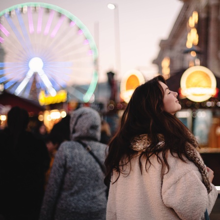 Weihnachtsmarkt in Dämmerung: Junge Frau mit erhobener Nase, lange Haare, Mantel, Rücken zur Kamera, viele Lichter unscharf im Hintergrund, Menschen, Riesenrad. Feierliche Stimmung.