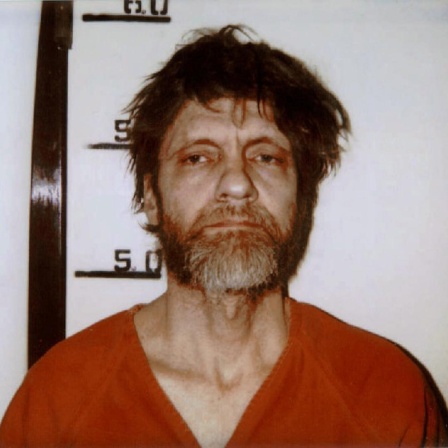 Mugshot von Ted Kaczynski in seinem orangeroten Gefängnisoverall. Sein Haar ist strubbelig und er schaut müde in die Kamera.