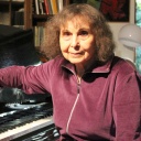 Die Komponistin Sofia Gubaidulina sitzt an ihrem schwarzen Flügel und lächelt.