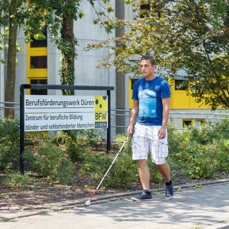 Ein Mann mit Sehbehinderung und einem weißen Stock geht auf einem Bürgersteig
